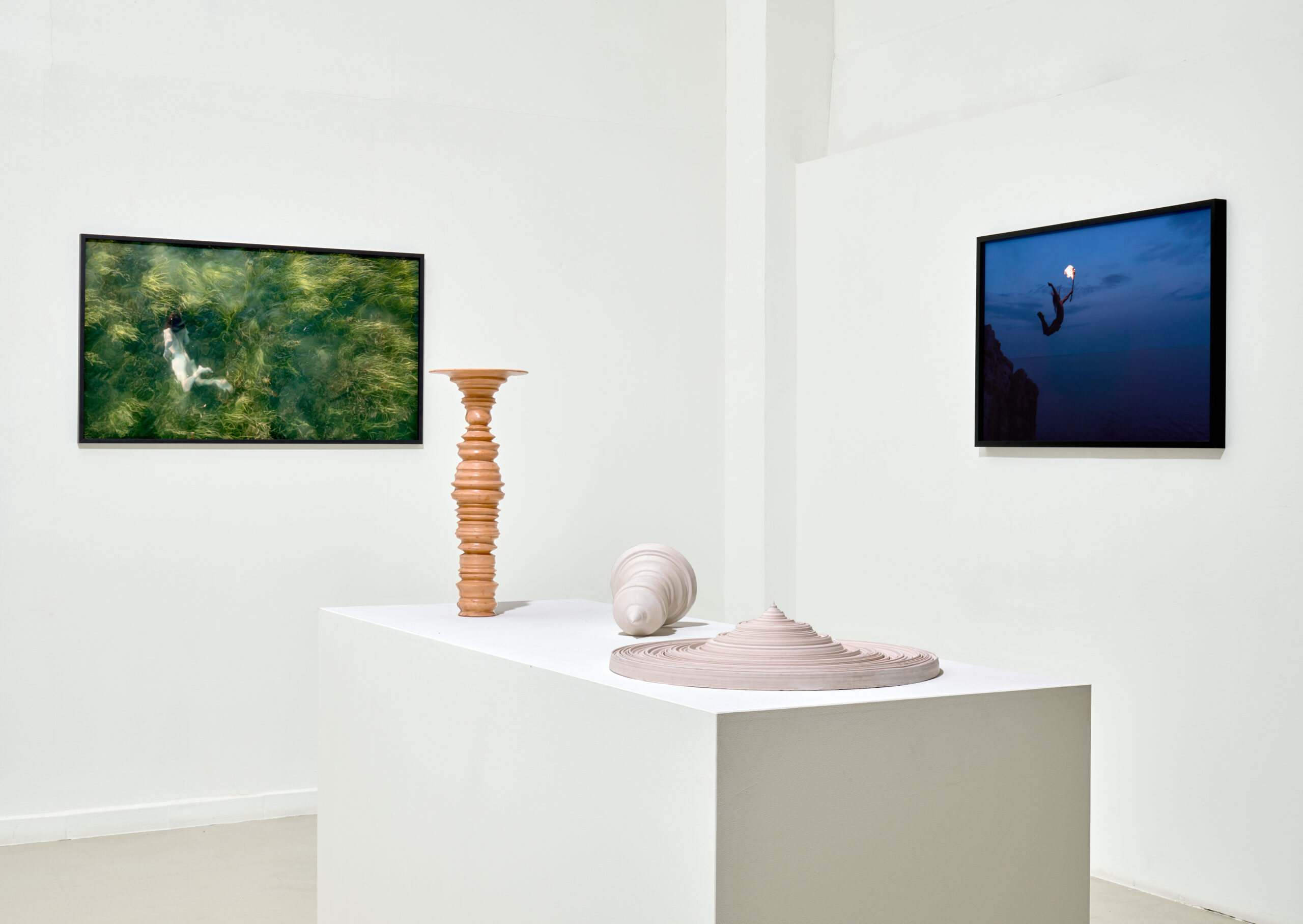 Vue d'exposition à la galerie Eva Vautier : deux photographies encadrées au mur et, sur un socle blanc, une colonne de bois tournée à côté d'une forme céramique claire
